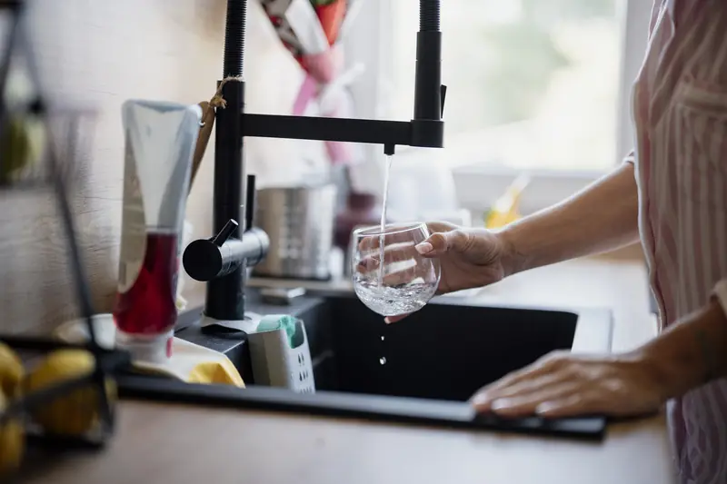 A hand filling a clear glass under a matt black mixer tap at a dark kitchen sink, with water splashing into the glass