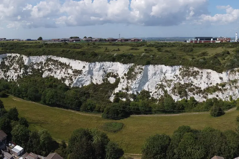 A long white wall of exposed chalk in the old Paulsgrove pit beneath the crest of Portsdown Hill, with scrub and a meadow in front and buildings along the skyline