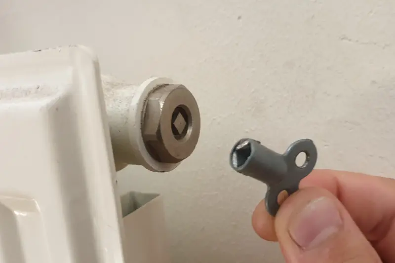 Fingers holding a small radiator key against the square bleed valve at the top corner of a white radiator