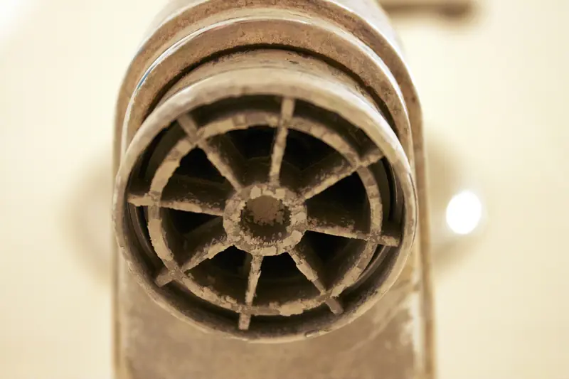 Close-up of a chrome tap spout whose round aerator mesh is crusted with pale grey limescale around its spokes