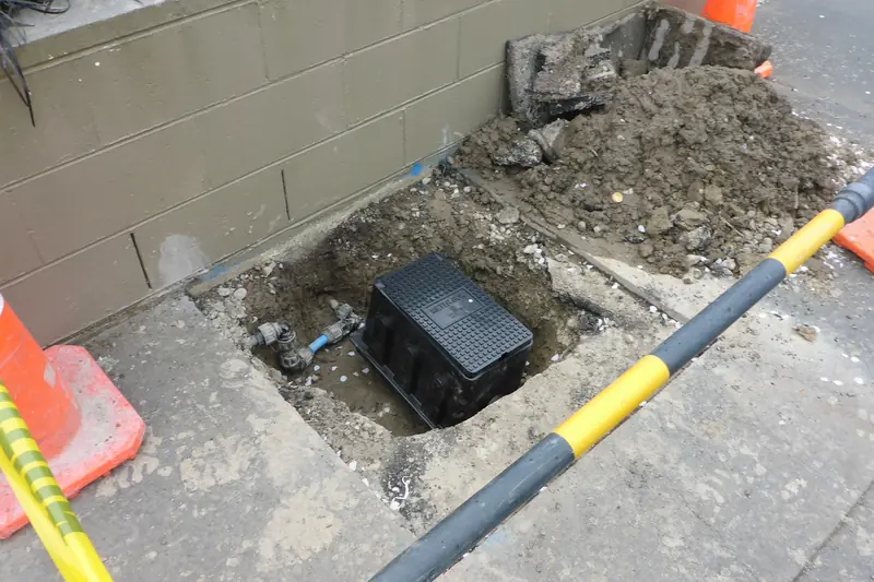 Water meter chamber dug open beside a paved footpath, with loose earth, a traffic cone and a black and yellow barrier pole